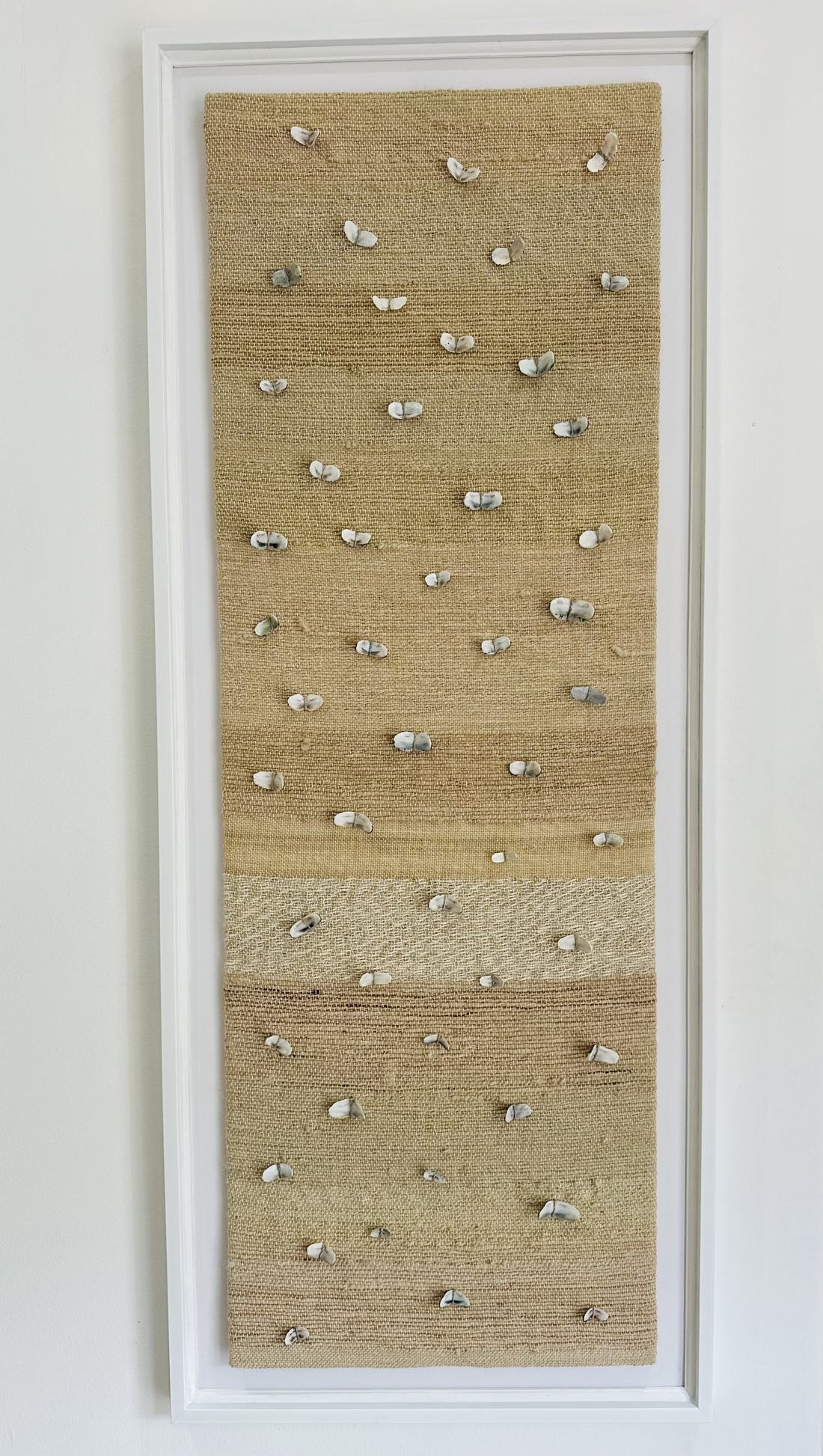 Tall narrow handwoven textile in warm sand tones with small pale shell shapes scattered across it like butterflies, framed in white canvas.